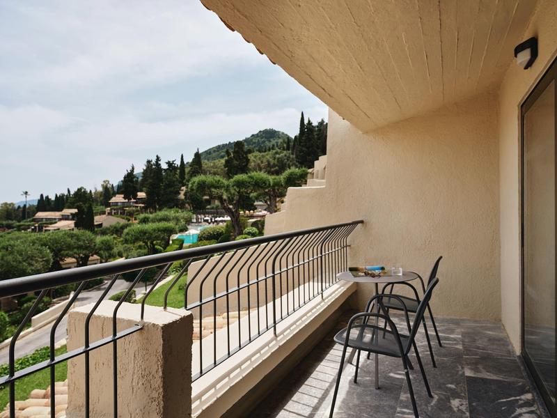 Balkon mit zwei schwarzen Stühlen, Tisch und Blick auf grüne Hügel unter bewölktem Himmel.