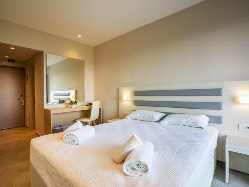 Helles Hotelzimmer mit Doppelbett, Nachttischen, Stuhl und großem Spiegel an der Wand.