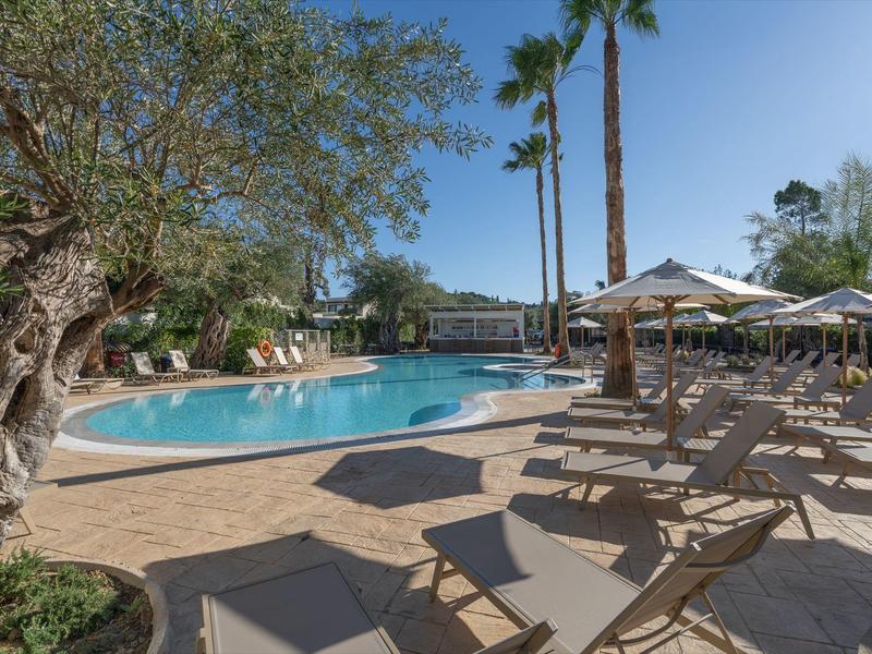 Ein Swimmingpoolbereich mit Sonnenliegen, Sonnenschirmen und Palmen an einem sonnigen Tag.