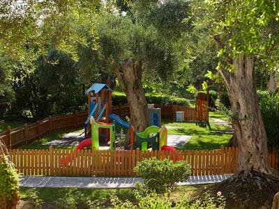 Kinderspielplatz mit Rutsche und Klettergerüst, umgeben von hohem Holzzaun und großen Bäumen.