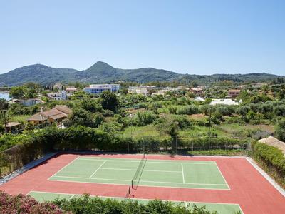 Tennisplatz mit rotem Belag vor grüner Landschaft und Bergen unter klarem blauem Himmel.