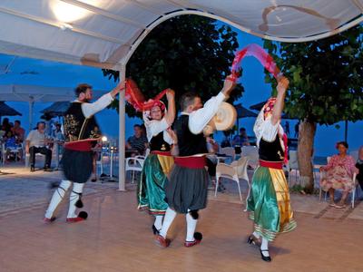 Vier Personen in traditioneller Kleidung tanzen im Freien unter einem Zelt mit Zuschauern.
