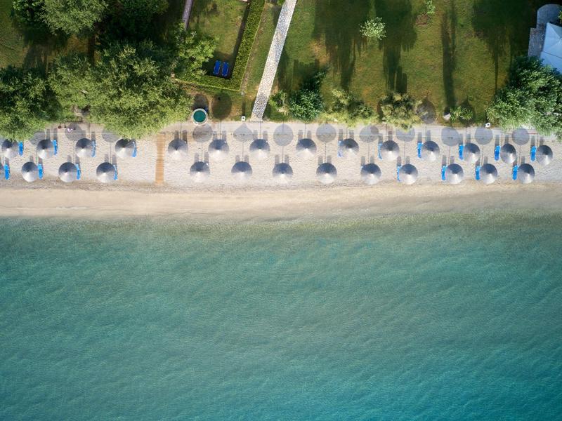 Luftaufnahme eines Strandes mit Sonnenschirmen und Liegestühlen neben klarem Wasser und Bäumen.