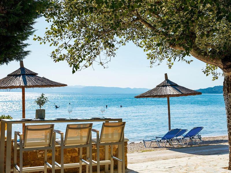 Strand mit Sonnenschirmen, Liegestühlen und einem Tisch mit Stühlen unter Bäumen am Meer.