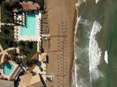 Strandhotel mit zwei Pools, Sonnenliegen, Sandstrand und Wellen am Meer in Vogelperspektive.
