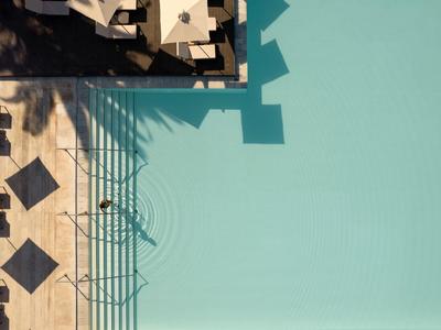 Schwimmbecken mit klar blauem Wasser, hellen Fliesen und Schatten von Zaun und Möbeln am Rand.