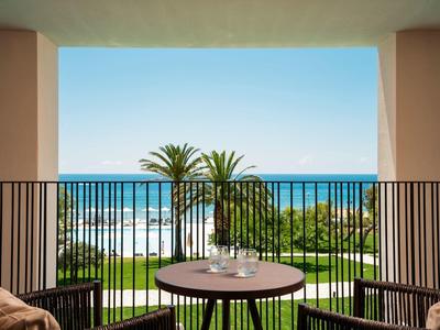 Blick von Balkon mit rundem Tisch und Stühlen auf blauen Ozean, grüne Palmen und Garten bei klarem Himmel.