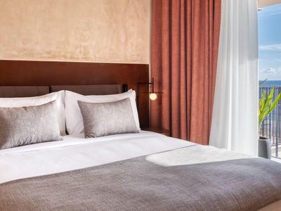 Helles Schlafzimmer mit großem Bett, rosafarbenen Vorhängen und Balkon mit Meerblick.