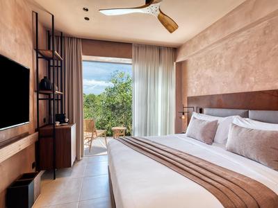 Helles Schlafzimmer mit großem Bett, Holzmöbeln und Blick auf grüne Pflanzen durch Glasfenster.