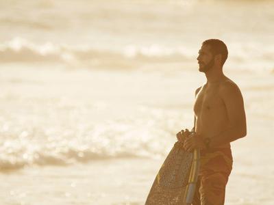 Mann steht am Strand mit Surfboard, sanfte Wellen und weiches Licht im Hintergrund.