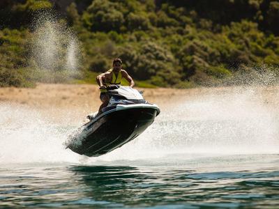 Person auf schwarzem Jetski springt über Wasser mit grünem bewaldetem Ufer im Hintergrund.