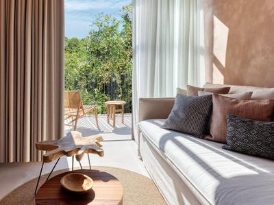 Helles Wohnzimmer mit weißem Sofa, Kissen, Holzstuhl und Glasfront zum begrünten Balkon.