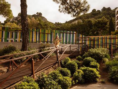 Holzsteg führt durch gepflegten Garten mit blühenden Sträuchern zu Hotelanlage am Hang.