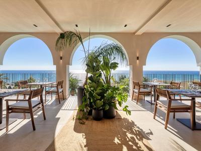 Offenes Restaurant mit Holztischen, Stühlen und Blick auf das Meer unter Arkaden.