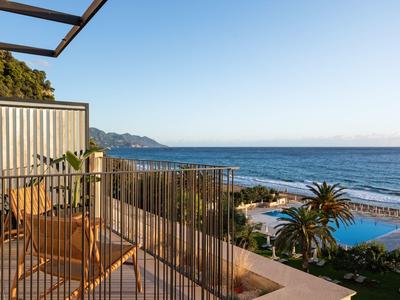 Balkon mit Holzstuhl, Metallgeländer, Blick auf Meer, Pool und Palmen unter klarem blauem Himmel.