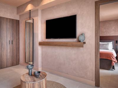 Modernes Hotelzimmer mit Fernseher, großem Spiegel, Holzmöbeln und Blick ins Schlafzimmer.