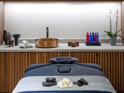Moderner Massagetisch mit grauer Decke, Steinen und Blumen vor Holzwand und beleuchtetem Regal.