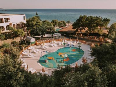 Runder Pool mit buntem Design, umgeben von Liegestühlen und Bäumen am Meer.
