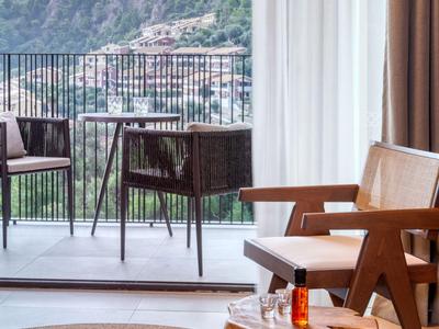 Balkon mit Stühlen und einem kleinen Tisch, Blick auf Berge und Stadt, helle Vorhänge im Zimmer.