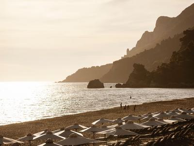 Sonnenuntergang über dem ruhigen Strand mit leeren Liegestühlen und Bergen im Hintergrund.