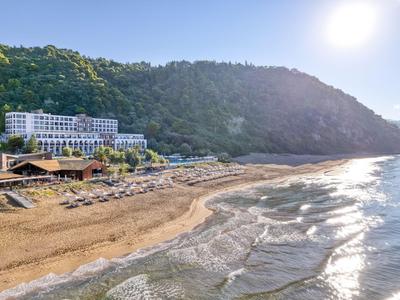 Hotelanlage am Strand mit Hügeln im Hintergrund und Sonne am klaren Himmel.