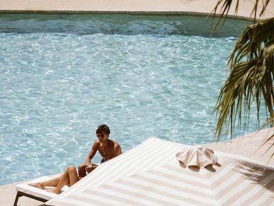 Eine Person entspannt sich auf einer Liege neben einem klaren Pool unter Palmen.
