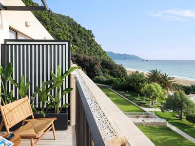 Balkon mit Stühlen und Pflanzen blickt auf einen Garten und das Meer bei klarem Himmel.