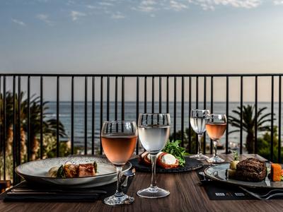 Blick auf gedeckten Tisch mit Glas Wein und Wasser, dahinter Meer und Palmen bei Sonnenuntergang.