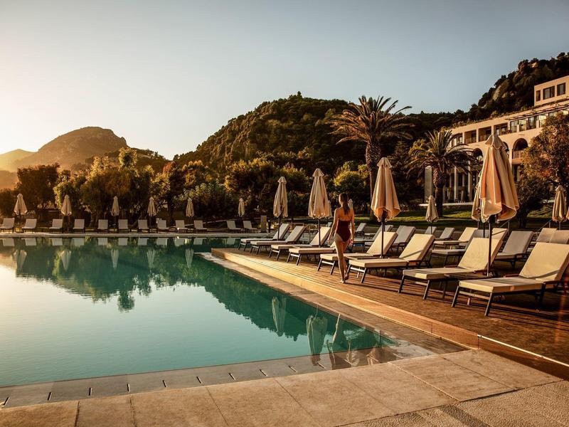 Großer Pool mit Liegestühlen und Sonnenschirmen bei Sonnenuntergang, umliegende grüne Hügel.