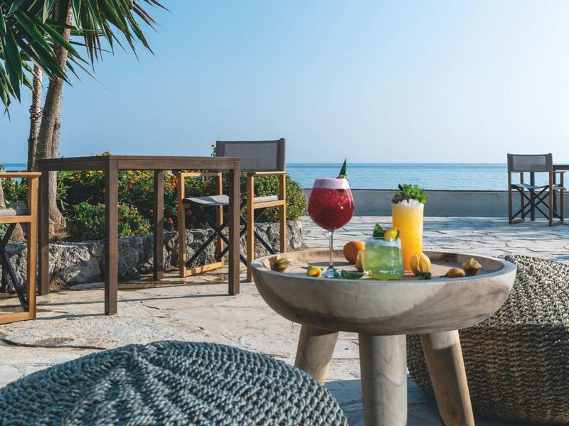 Strandbar mit Holzmöbeln, zwei Getränken auf runden Tischen und Palmen vor klarem Himmel.