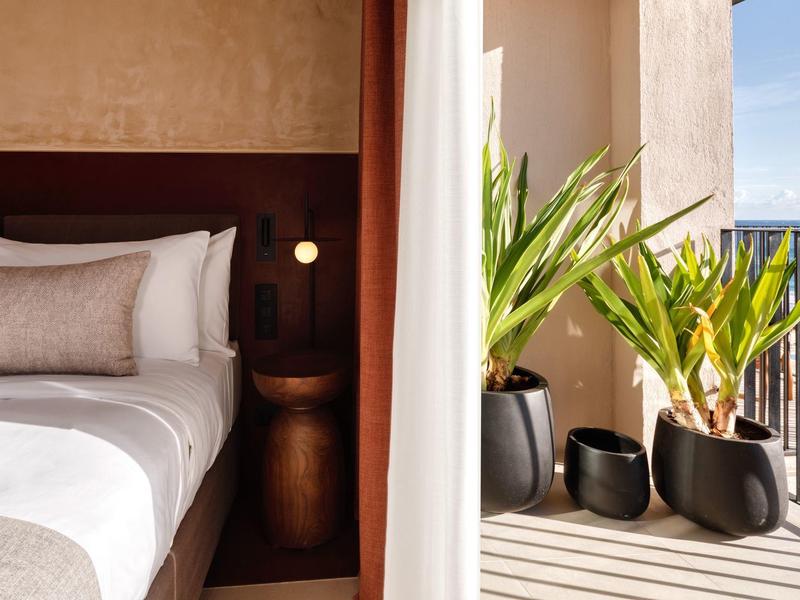 Helles Schlafzimmer mit großem Bett, braunen Holzmöbeln und Pflanzen auf Balkon mit Blick auf Meer.