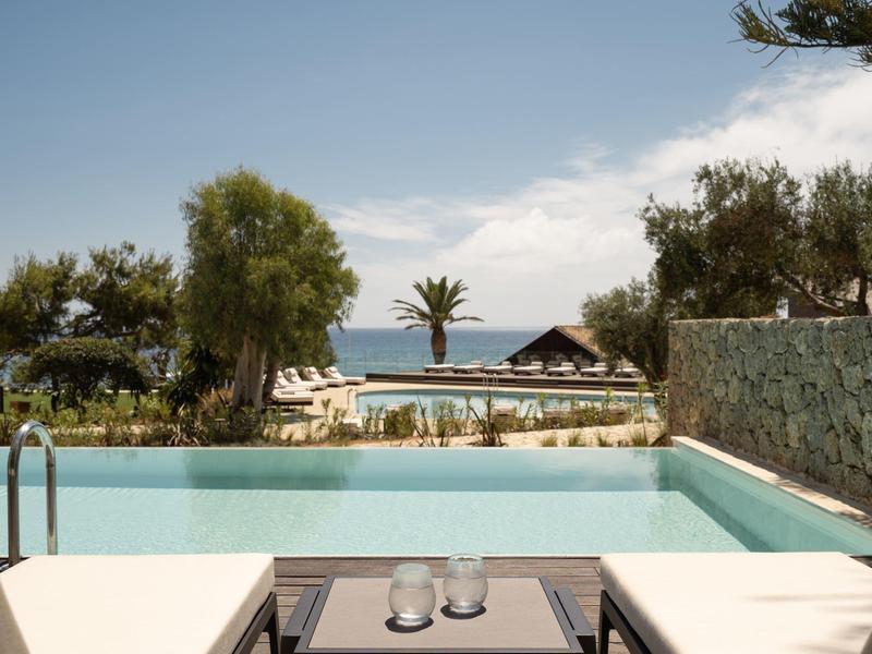 Pool mit klarem Wasser, umgeben von Pflanzen, Sitzbereich und Meerblick unter blauem Himmel.