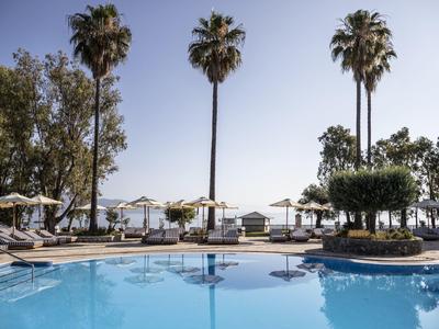 Blauer Pool mit Liegestühlen, Sonnenschirmen und hohen Palmen vor klarem Himmel am Meer.