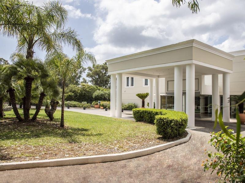 Eingang eines Gebäudes mit weißem Vordach, umgeben von Pflanzen und Palmen bei bewölktem Himmel.