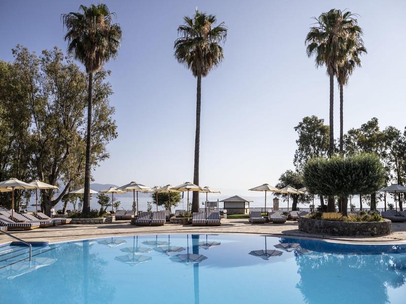 Blauer Pool mit Liegestühlen, Sonnenschirmen und hohen Palmen vor klarem Himmel am Meer.