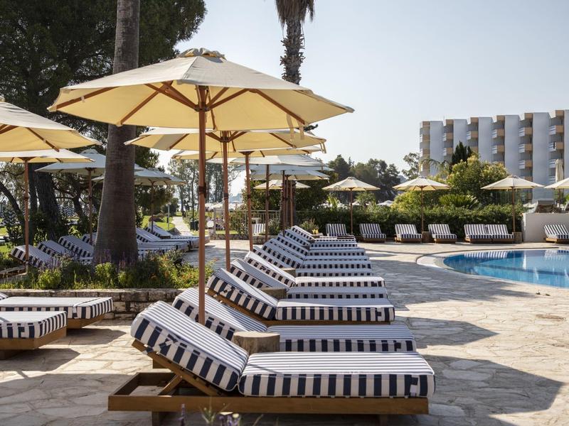 Poolbereich mit Liegestühlen, weißen Sonnenschirmen und modernen Gebäuden im Hintergrund.