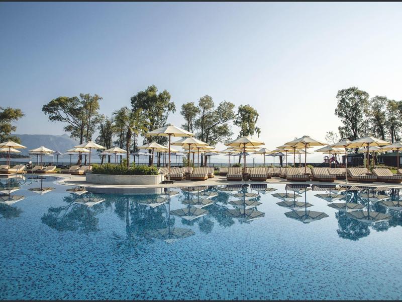 Großer Pool mit Sonnenschirmen und Bäumen neben dem Meer bei klarem Himmel.