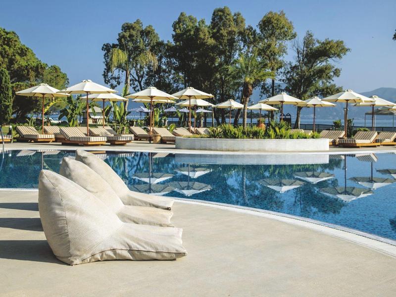 Weißer Sitzsack am Rande eines Pools mit Sonnenliegen und Sonnenschirmen im Hintergrund.