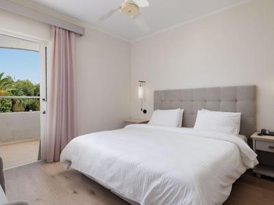 Helles Hotelzimmer mit Doppelbett, Nachttischen und Zugang zum Balkon mit Palmenblick.