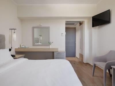 Schlichtes Schlafzimmer mit weißer Bettwäsche, grauem Sessel, integriertem Schreibtisch und Wandfernseher.