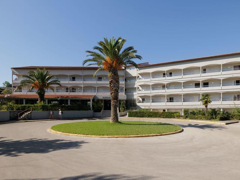 Mehrstöckiges weißes Gebäude mit Balkonen, umgeben von Palmen und einem Kreisverkehr im Vordergrund.