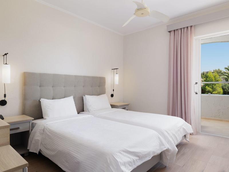 Helles Schlafzimmer mit zwei Einzelbetten, grauem Kopfteil, weißen Bettwäsche und Balkon mit Palmenblick.