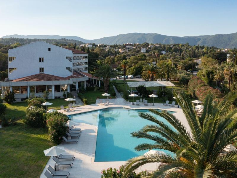 Großes weißes Haus mit rotem Dach und blauem Pool, umgeben von grünen Gärten und Bergen im Hintergrund.