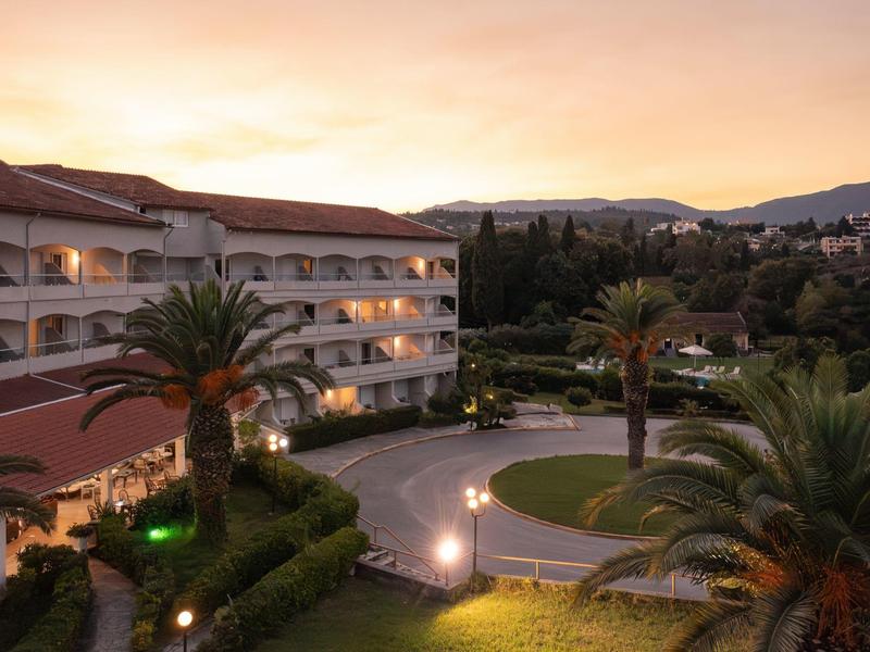 Hotel mit warm beleuchteten Balkonen, Palmen und Bergen im Hintergrund bei Sonnenuntergang.