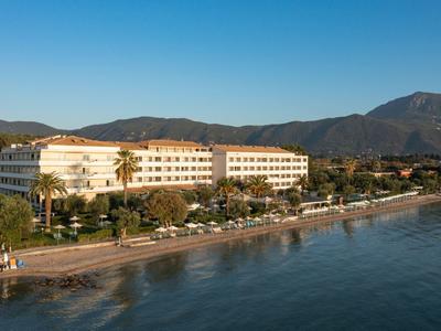 Großes Hotel mit roten Dächern am Meer, umgeben von Palmen und Bergen im Hintergrund.