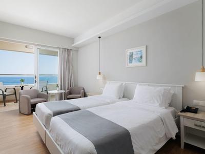 Doppelzimmer mit zwei Betten, grauen Decken, Holzfußboden, grauem Sessel und Meerblick durch große Fenster.