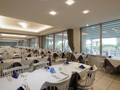 Großer Speisesaal mit weißen Tischen, blauen Servietten, vielen Stühlen und bodentiefen Fenstern.