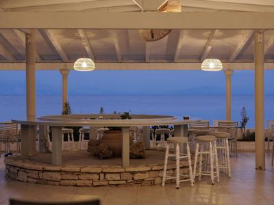 Offene Terrasse mit Bar, hellen Stühlen und Tischen, Blick auf ruhiges blaues Meer bei Abenddämmerung.