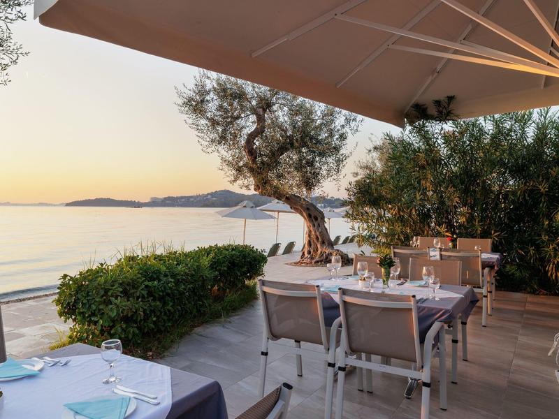 Terrasse mit gedeckten Tischen, Olivenbäumen, Blick auf Meer und Hügel bei Sonnenuntergang.