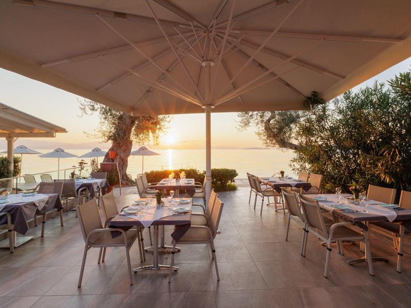 Terrassenrestaurant mit Tischen, Stühlen und Sonnenschirmen bei Sonnenuntergang am Meer.